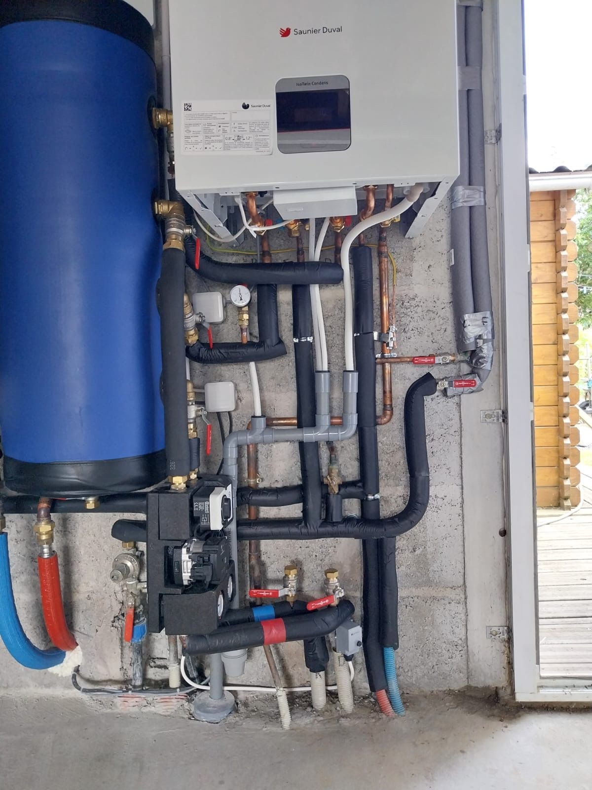 Chaufferie avec réservoir bleu, groupe électrogène blanc et tuyaux et vannes apparents sur un mur en béton.