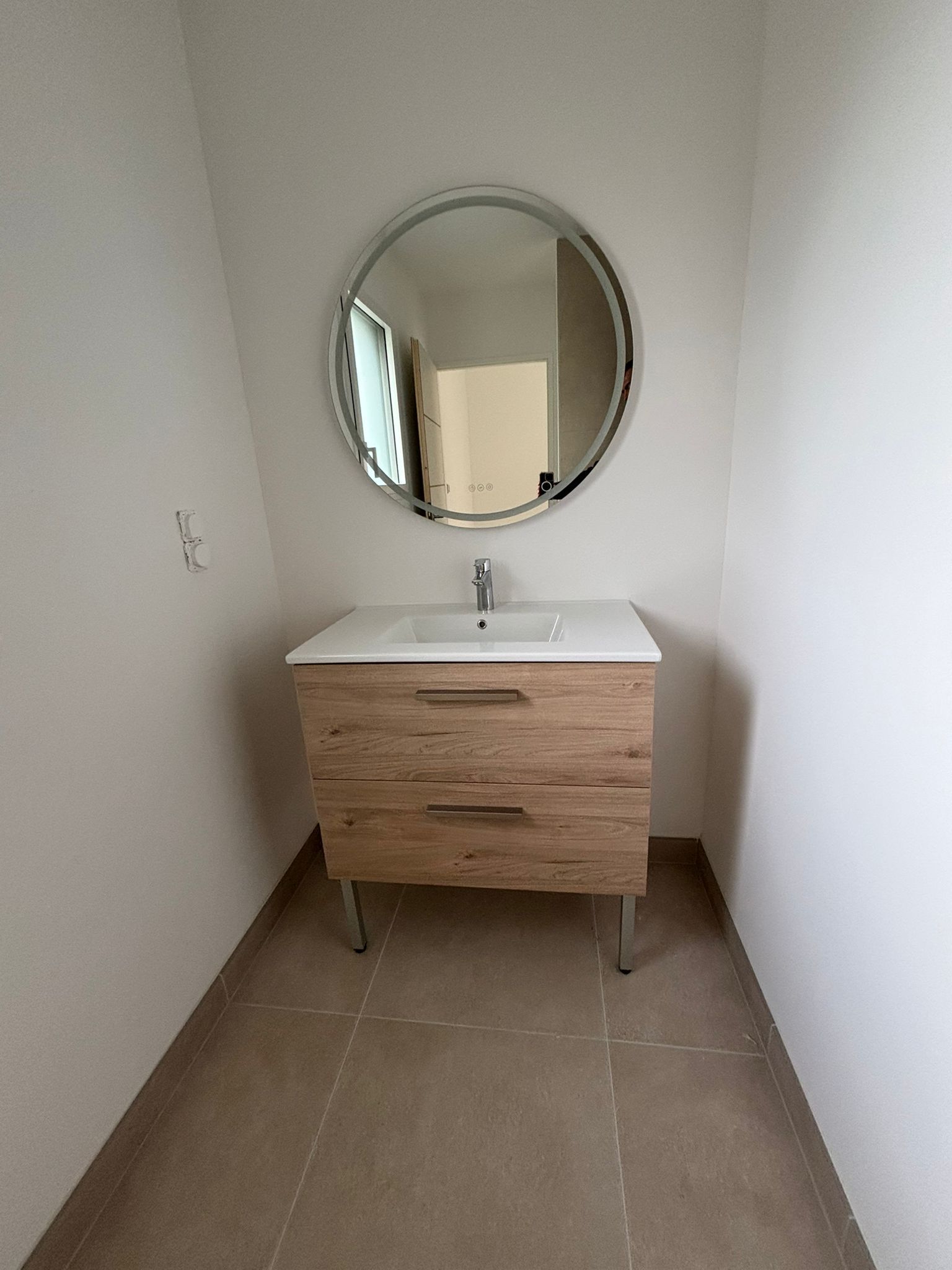 Meuble-lavabo étroit avec miroir rond, lavabo blanc et armoire en bois dans une petite niche carrelée.