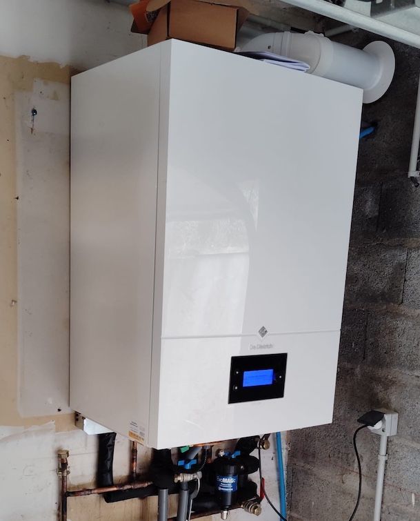 Chaudière murale blanche avec un petit écran bleu dans une buanderie, tuyaux visibles en dessous.