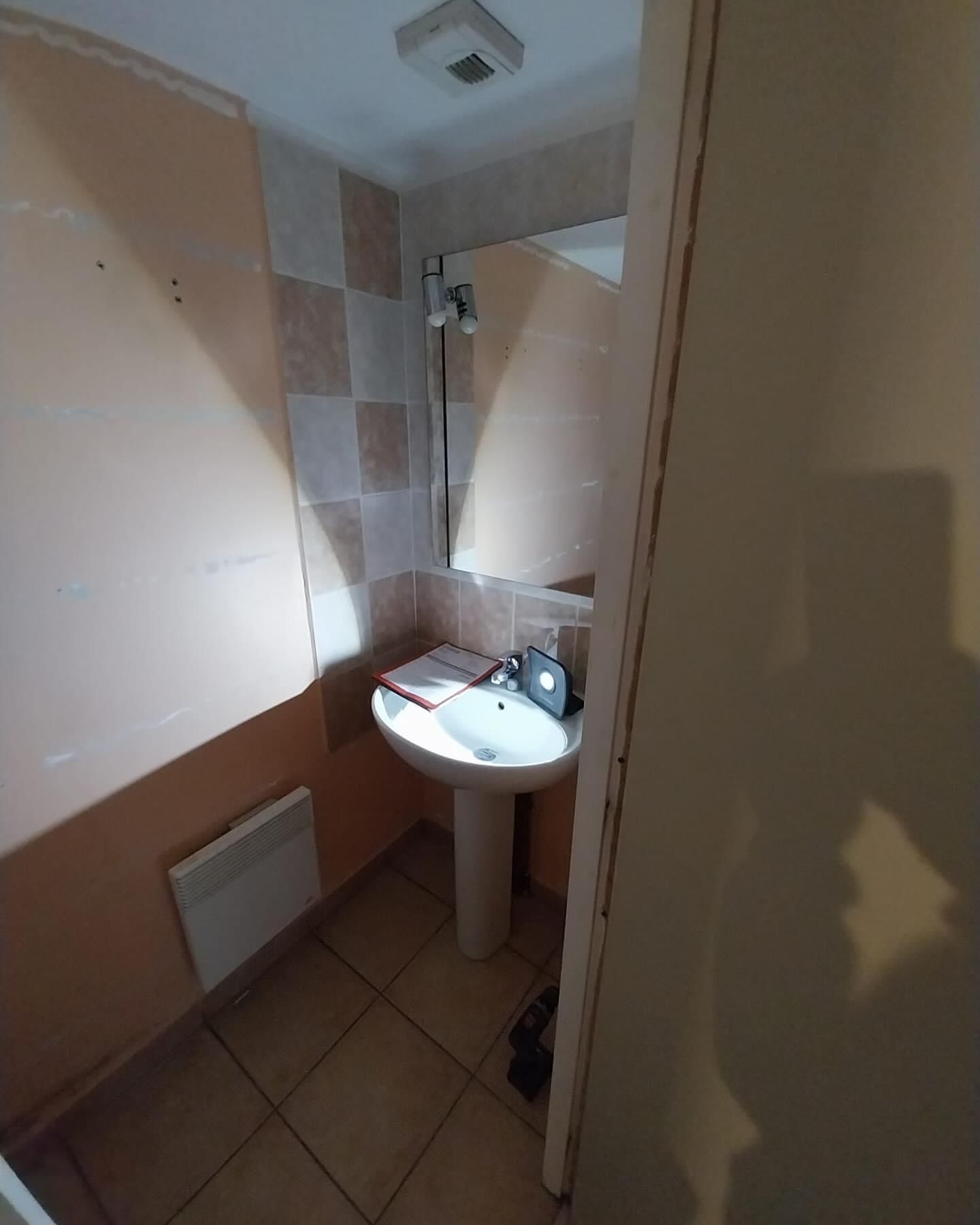 Petit coin lavabo dans une salle de bain, avec murs beiges, miroir et une serviette ou un sac posé sur le lavabo.