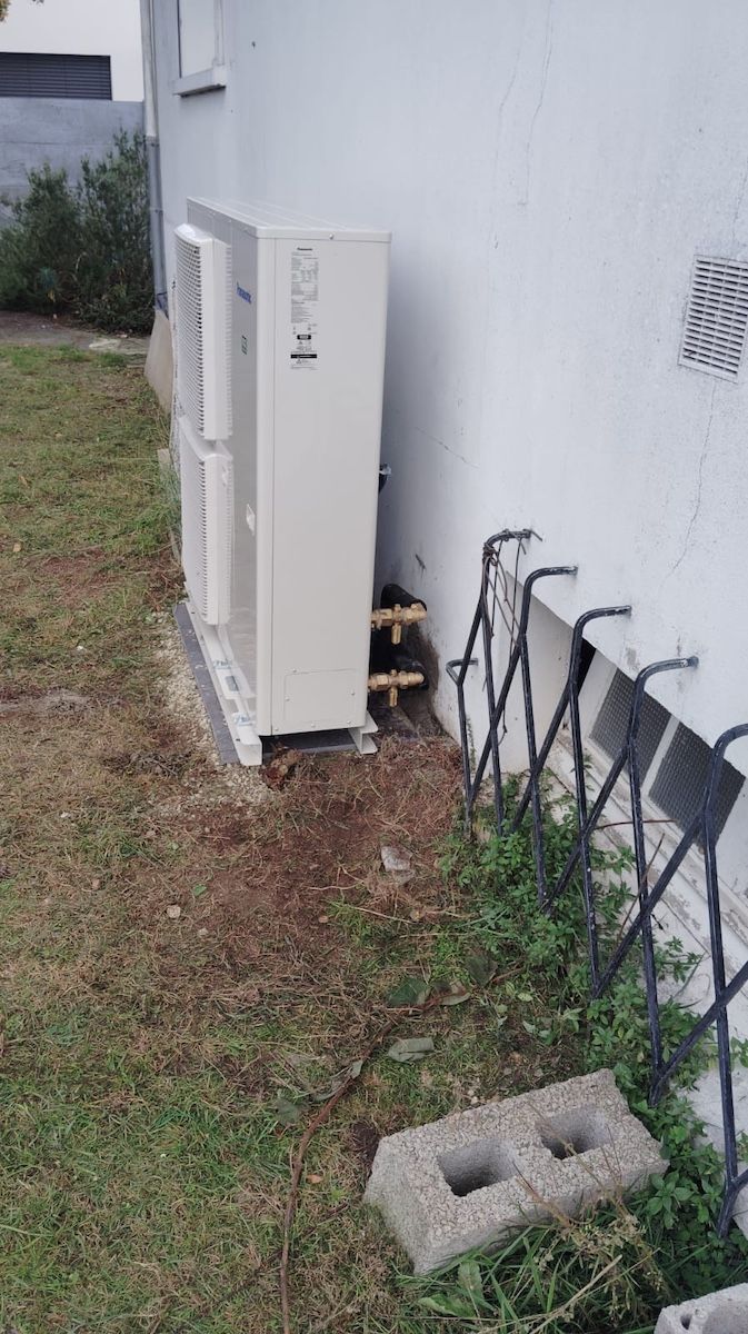 Des unités techniques extérieures et des câbles jouxtent un mur blanc, devant lequel se trouvent une cour gazonnée et un bloc de béton.