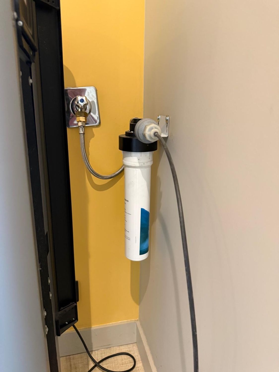 Filtre à eau mural avec tuyaux, à côté d'un mur jaune et d'un coin beige.