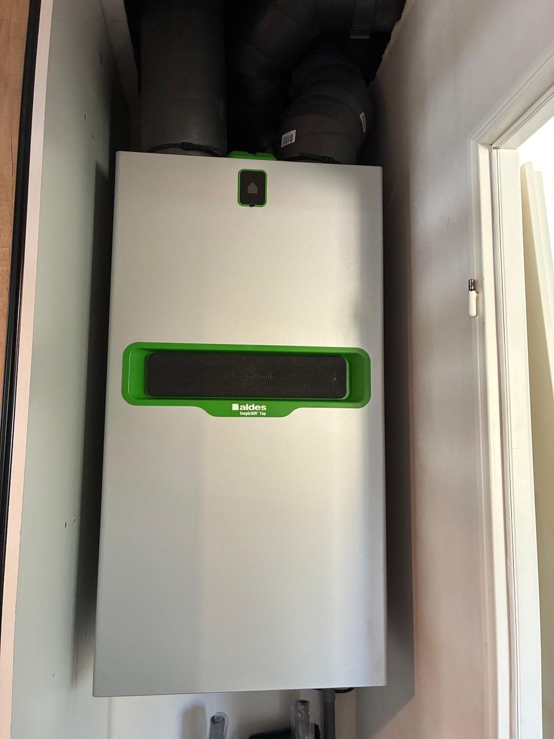 Appareil électroménager blanc fixé au mur avec des garnitures vertes dans un placard technique étroit, tuyaux de ventilation au-dessus.