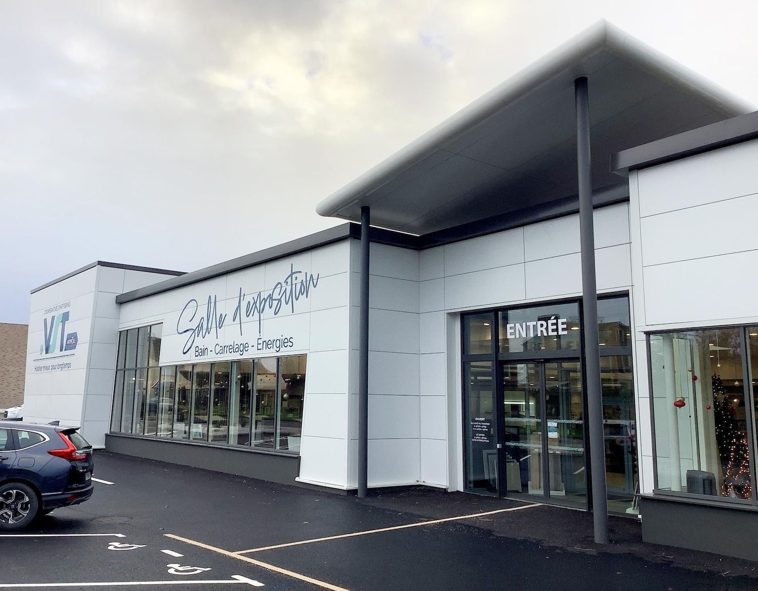 Façade moderne avec de grandes vitrines, façade blanche, toit noir et voitures garées à l'extérieur.