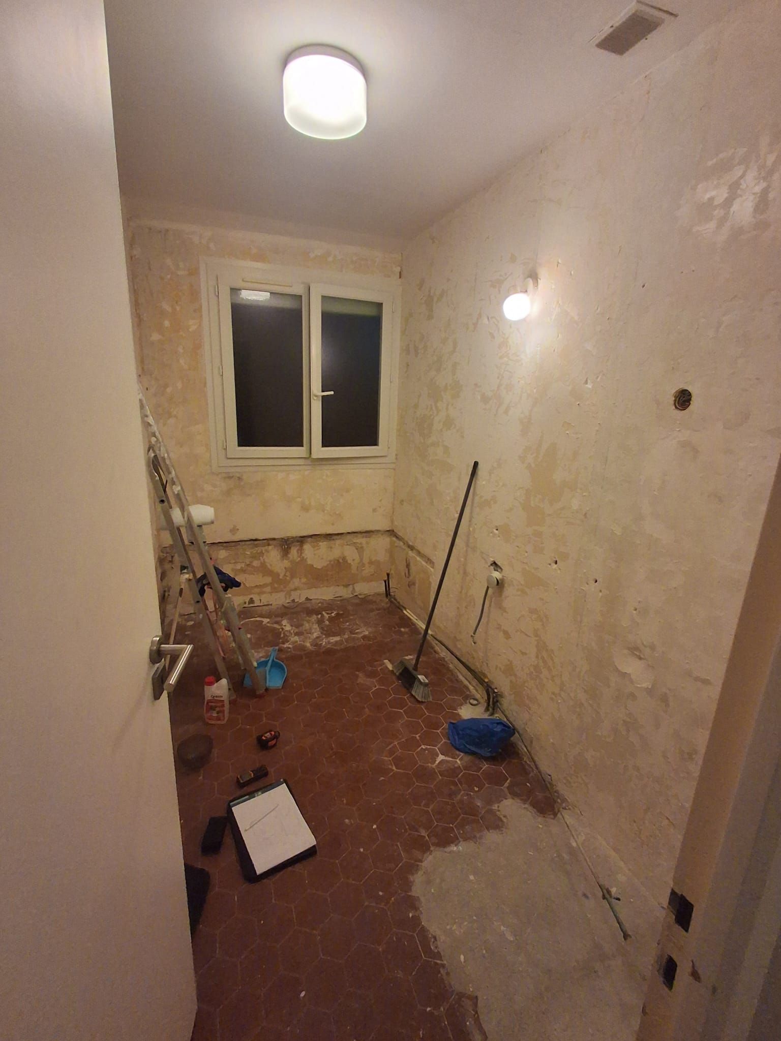 Petite pièce inachevée aux murs nus, avec des outils et du matériel de peinture éparpillés sur le sol près d'une fenêtre.