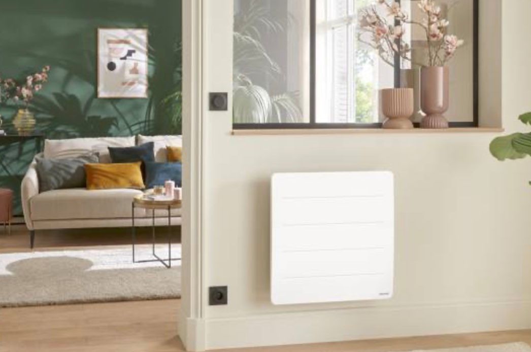 Salon avec une grille d'aération ou un radiateur blanc fixé au mur beige, à côté d'une fenêtre.