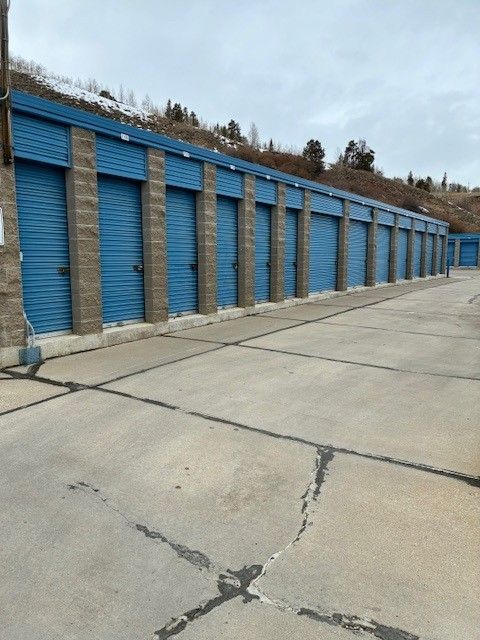 Exterior view of blue storage units lined up along a concrete lot, with a hillside in the background.