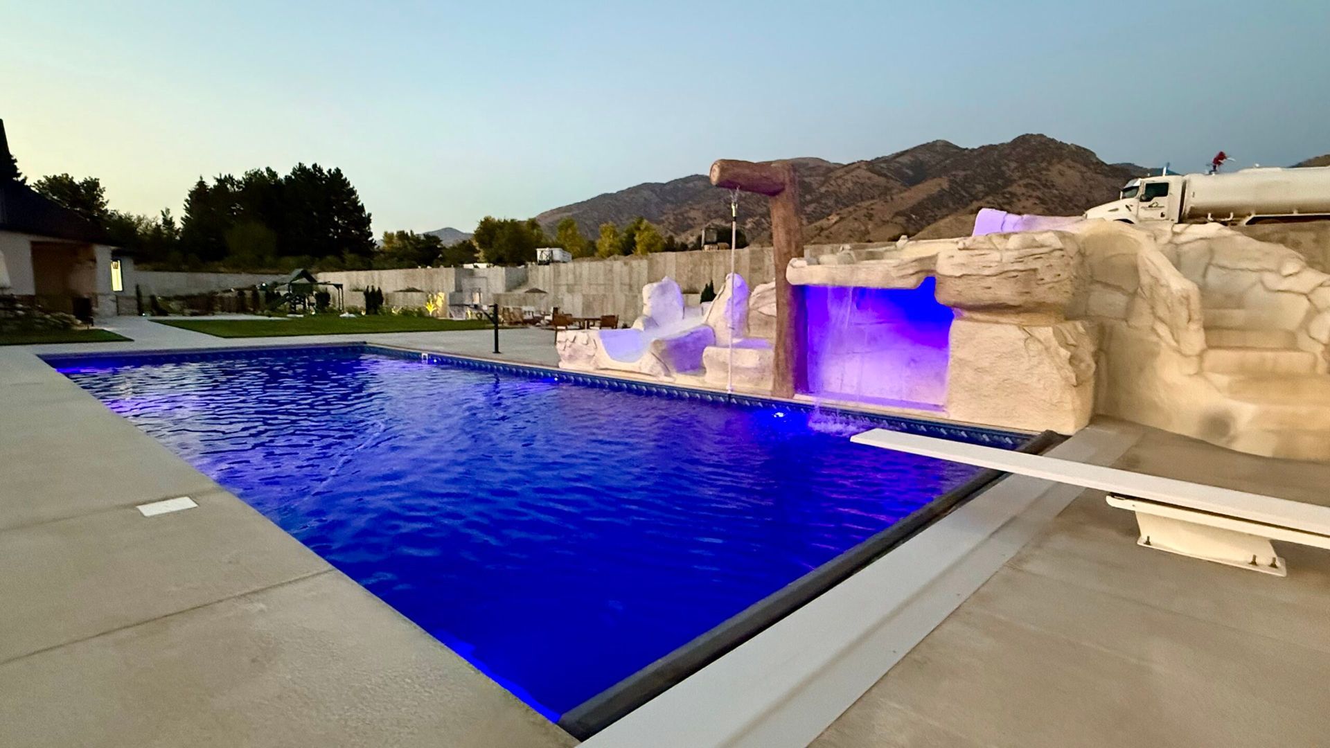 Outdoor pool with waterfall feature, diving board, and illuminated blue water, set against a mountainous background.