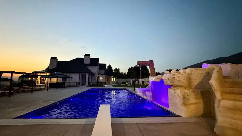 Pool at dusk with a diving board, water slide, and house in the background; purple pool lights.