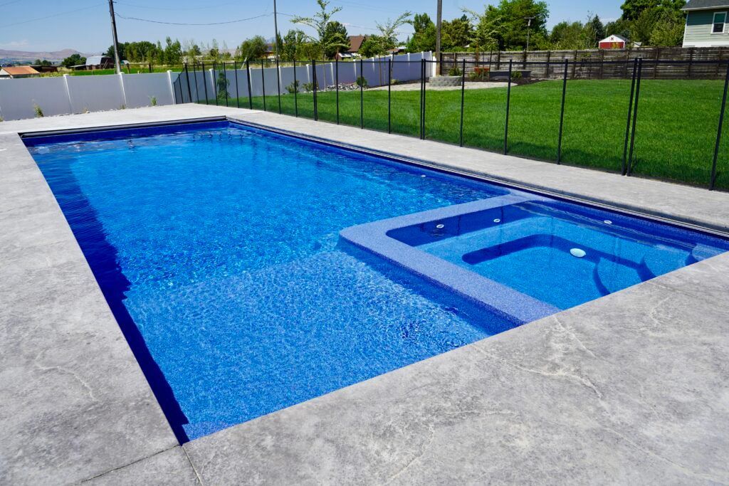 Blue rectangular swimming pool and attached jacuzzi, concrete patio, green lawn, black fence, sunny day.