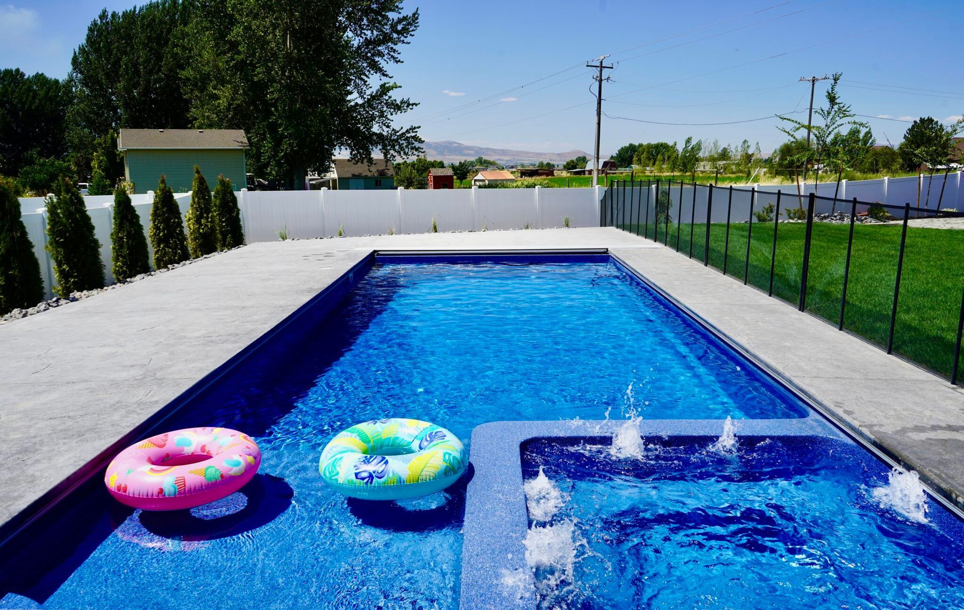 Rectangular pool with inflatables. Blue water, concrete deck, fountains. Sunny day, residential backyard setting.