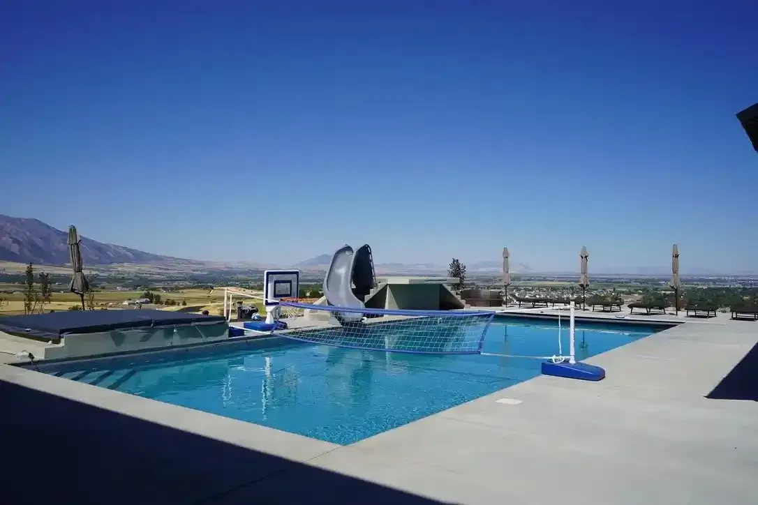Swimming pool with a water slide, basketball hoop, and mountains in the background under a blue sky.