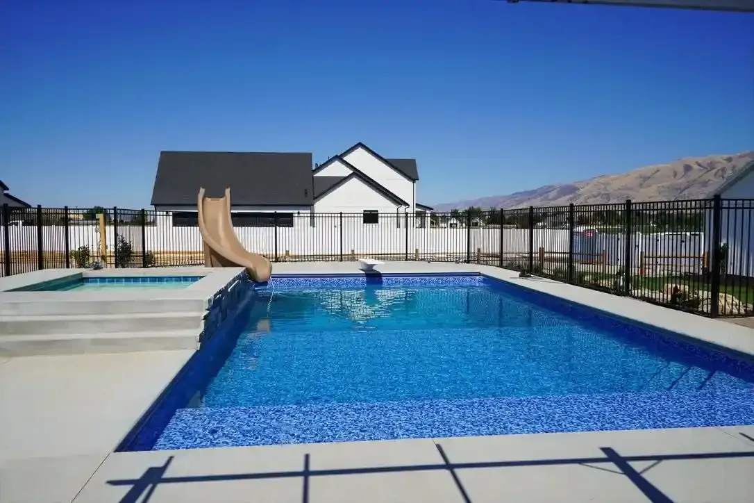 Backyard pool with slide, spa, and house against a blue sky, surrounded by a black fence.