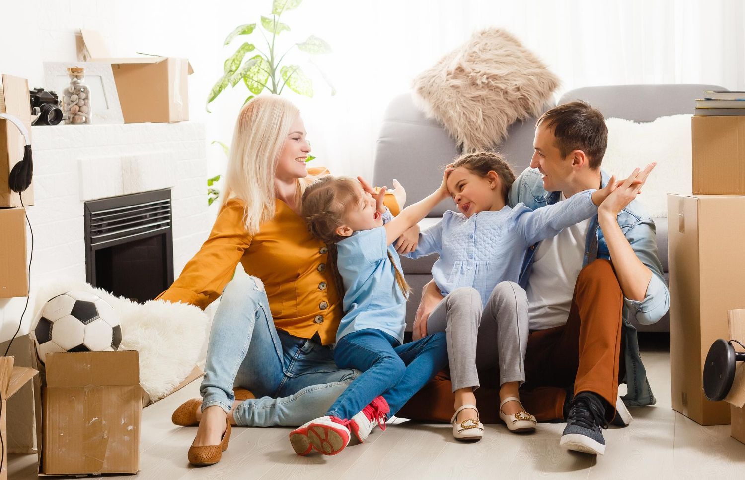A family is sitting on the floor in a living room surrounded by cardboard boxes.