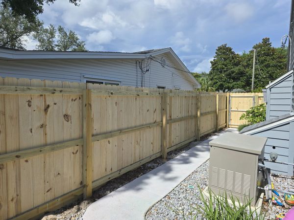 A new wooden fence runs along a concrete path next to a tan outdoor generator unit near a house.