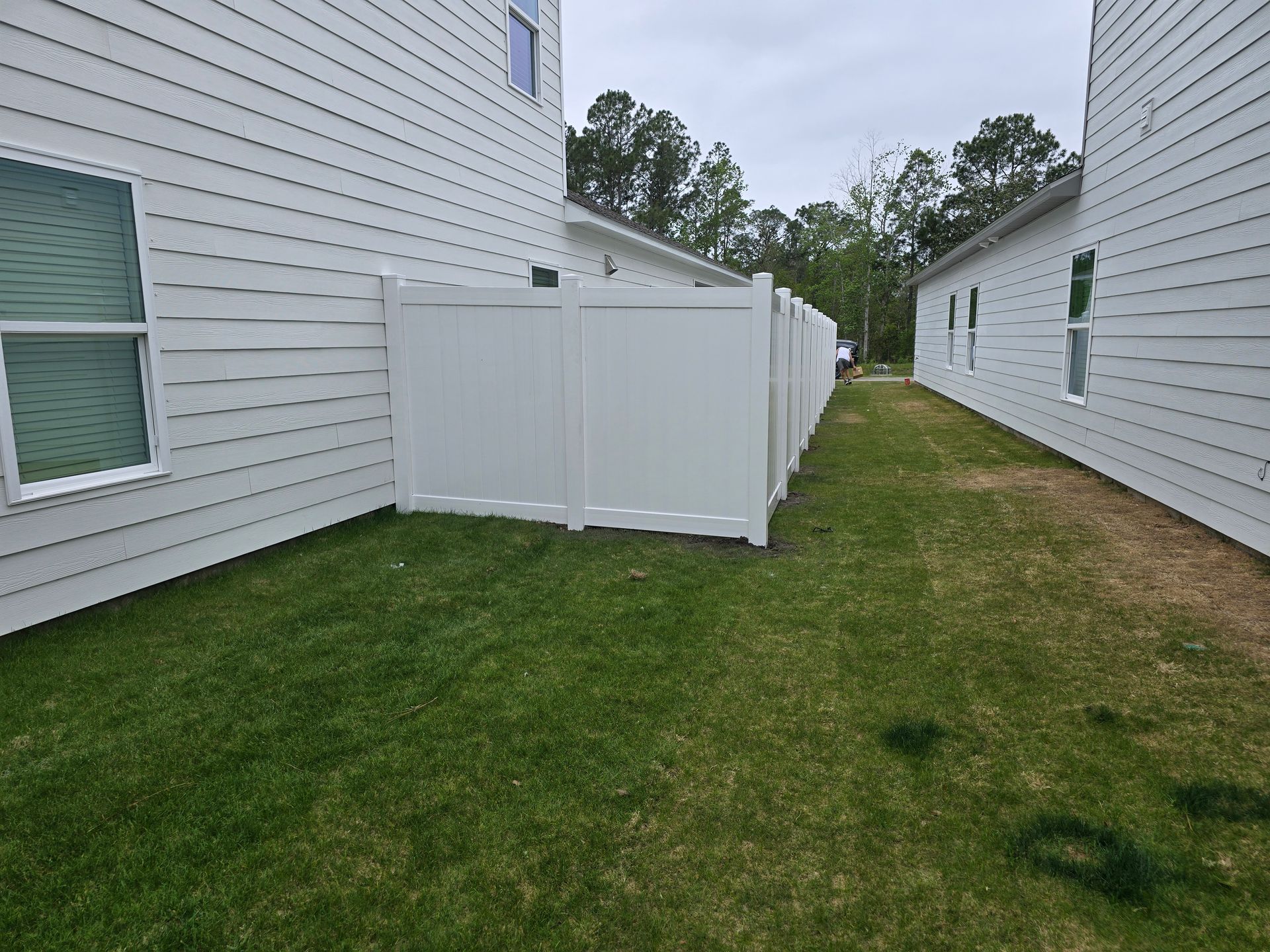 A narrow grassy path between two white-sided residential buildings, blocked by a tall white privacy fence.