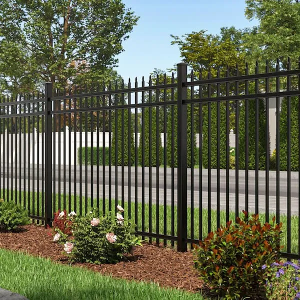 A black metal picket fence with pointed tops, situated in a front yard with green grass, mulch, and landscaping shrubs.