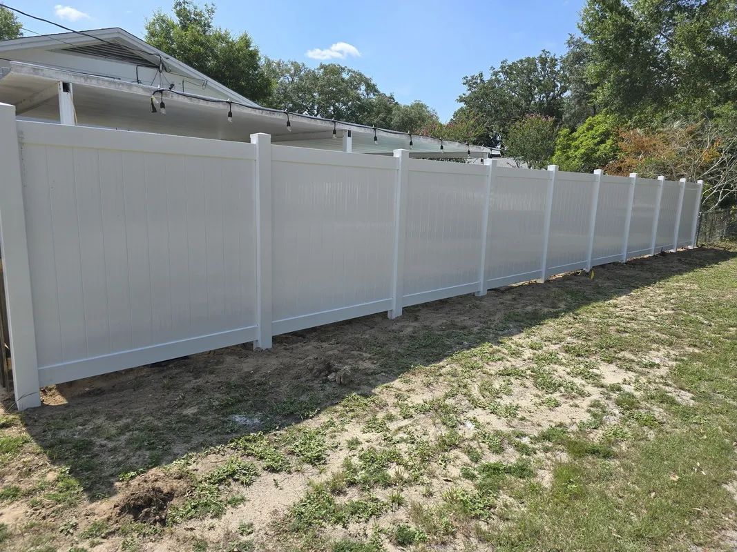 A long, white, solid vinyl privacy fence stands in a grassy yard, partially obscuring a house in the background.