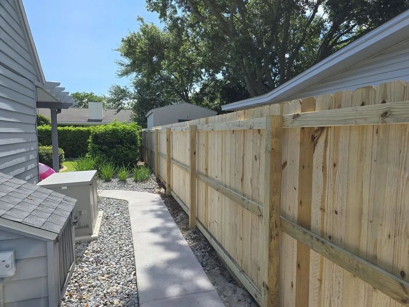 A wooden privacy fence borders a walkway between a house and a gravel area with a utility unit.