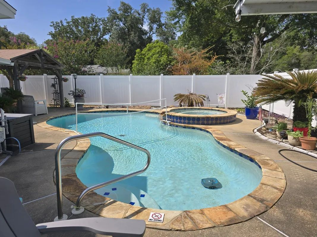 An outdoor swimming pool with a connected hot tub, surrounded by a concrete patio and white vinyl fence on a sunny day.
