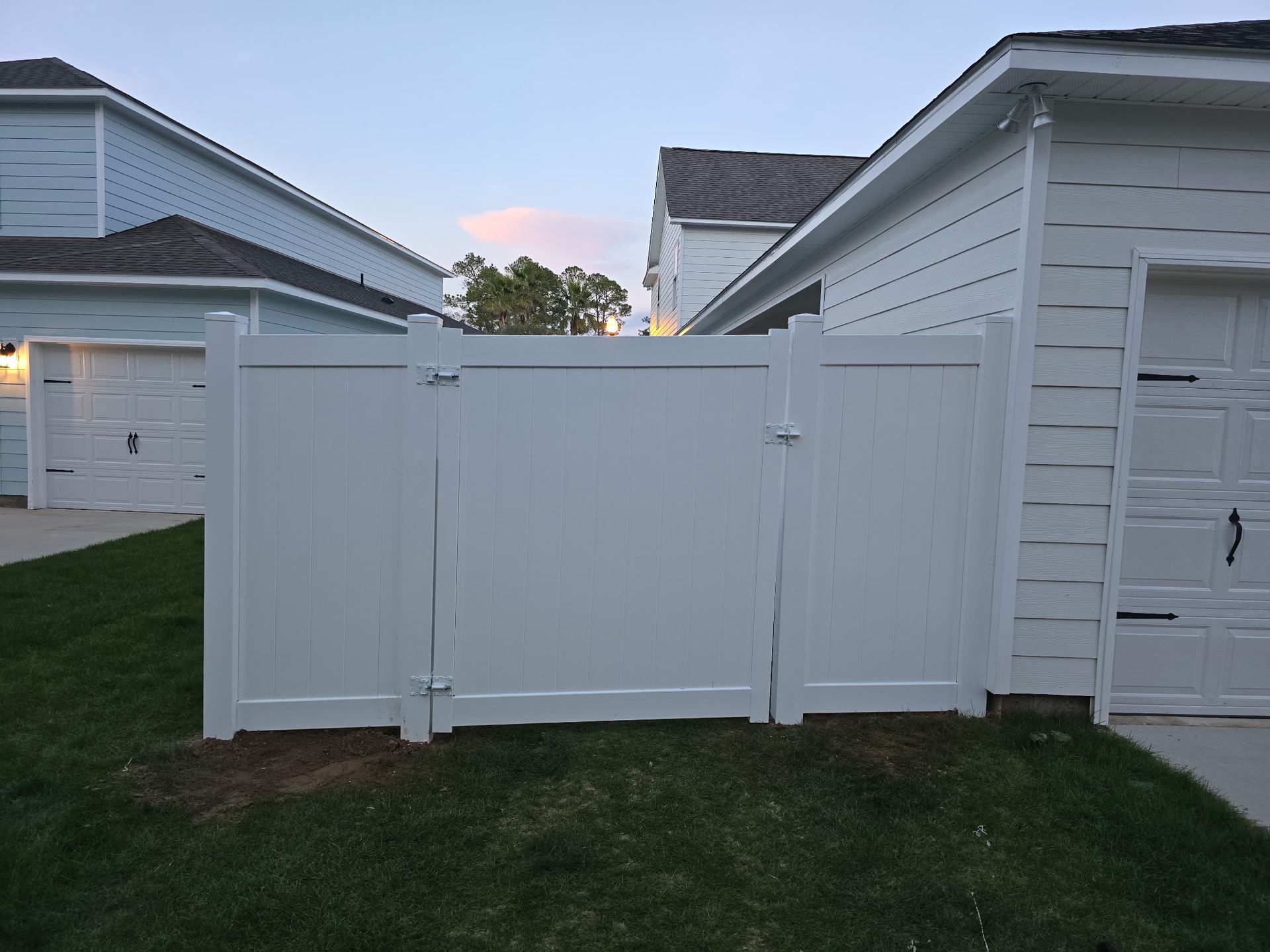 A white vinyl privacy fence with a central gate sits between two houses with white siding on a grassy lawn.