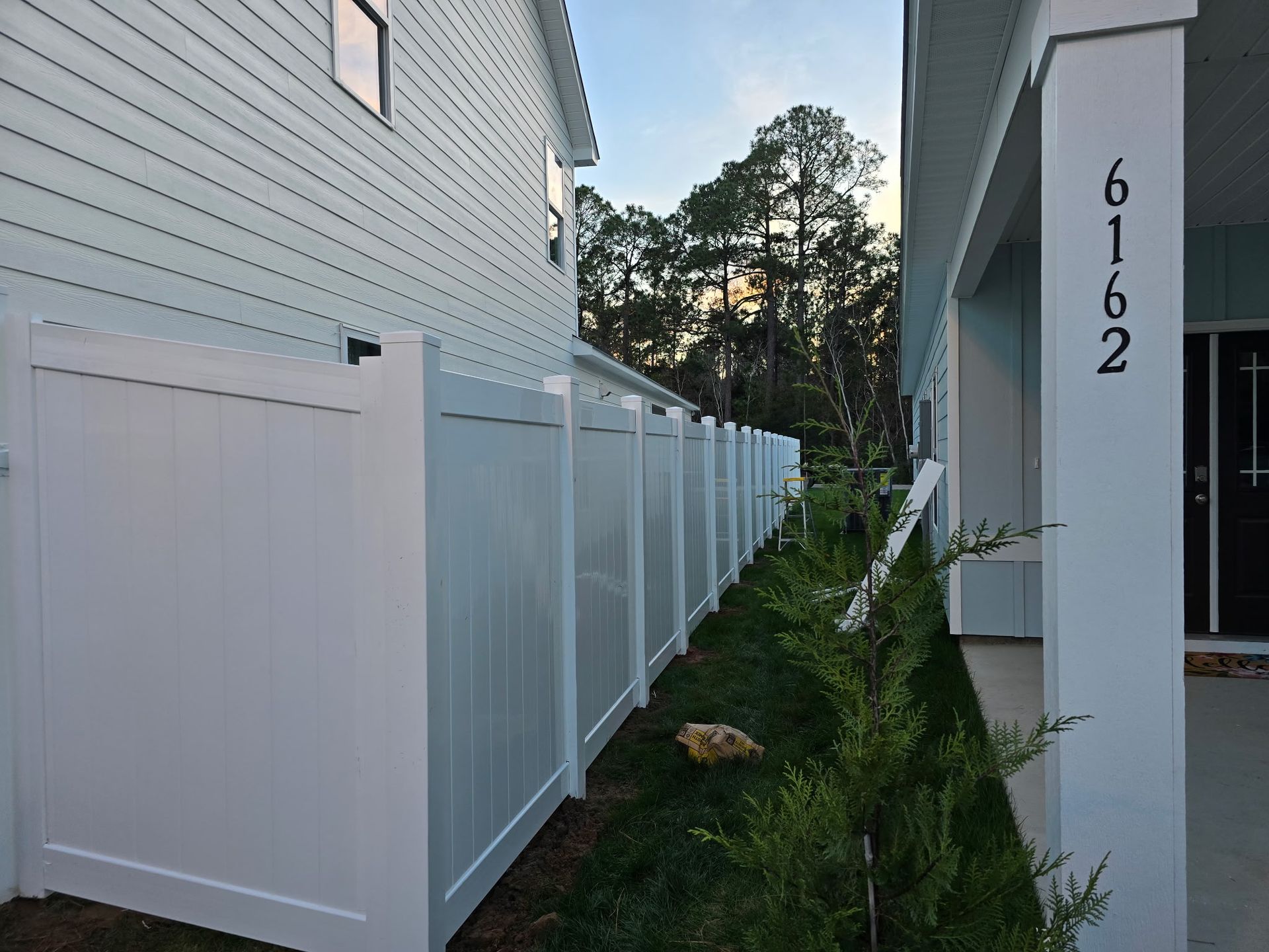 A side view of a white vinyl privacy fence running along the side of a light blue house with address number 6162.