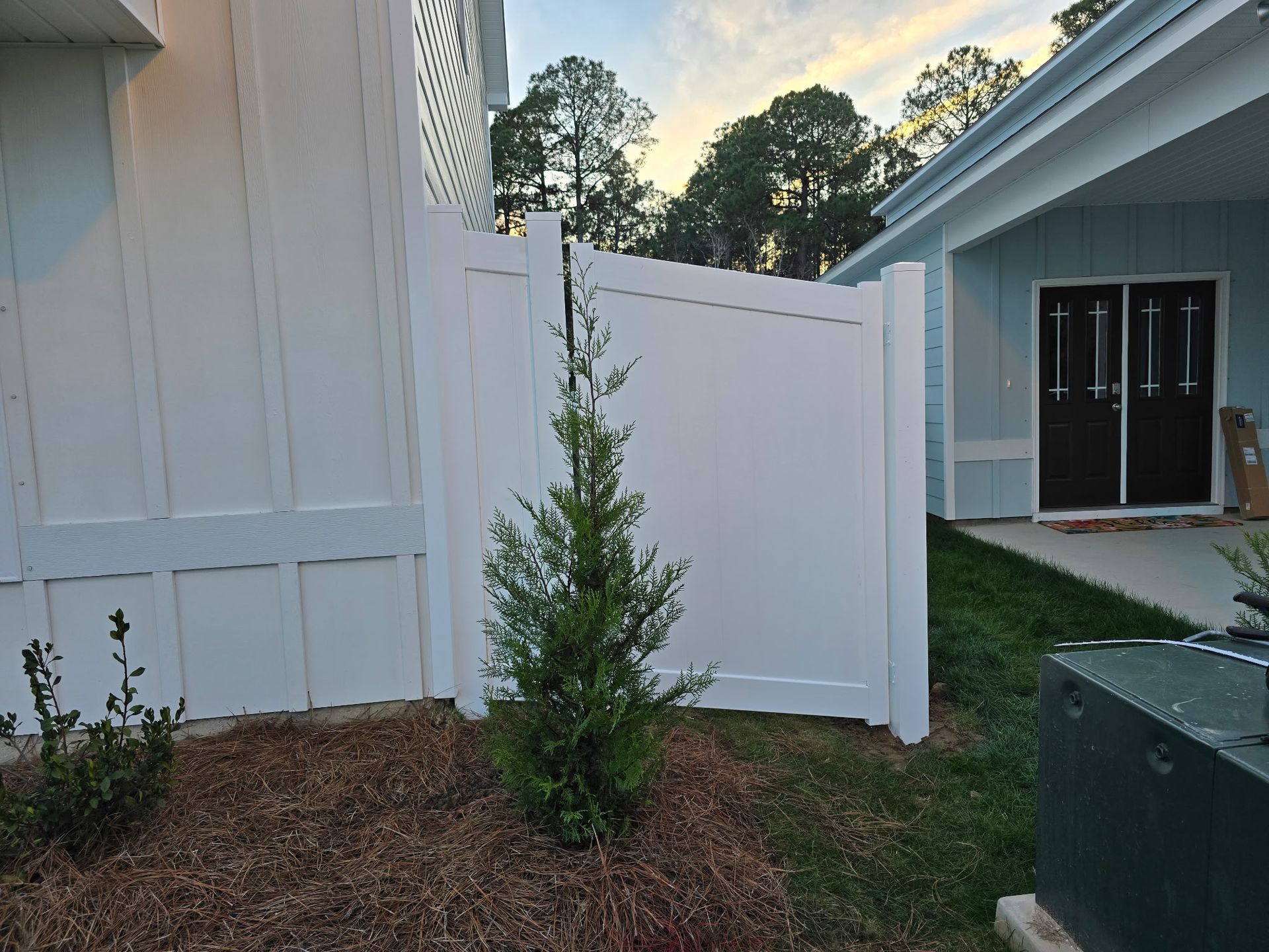A white vinyl fence stands between a house with white siding and a blue building with dark double doors.