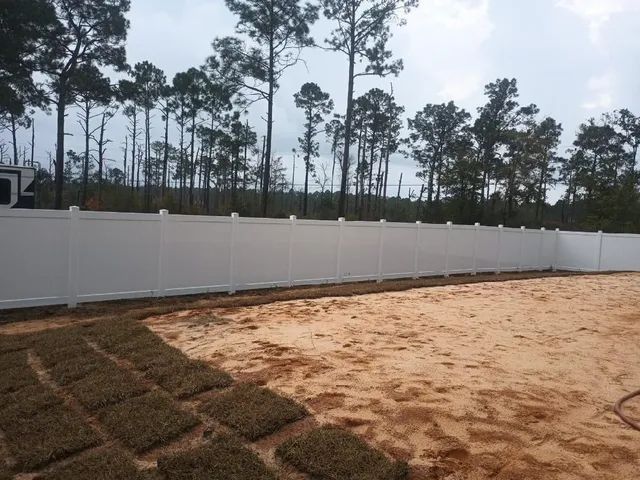 A tall, white vinyl privacy fence lines a sandy backyard partially covered with new patches of sod grass.