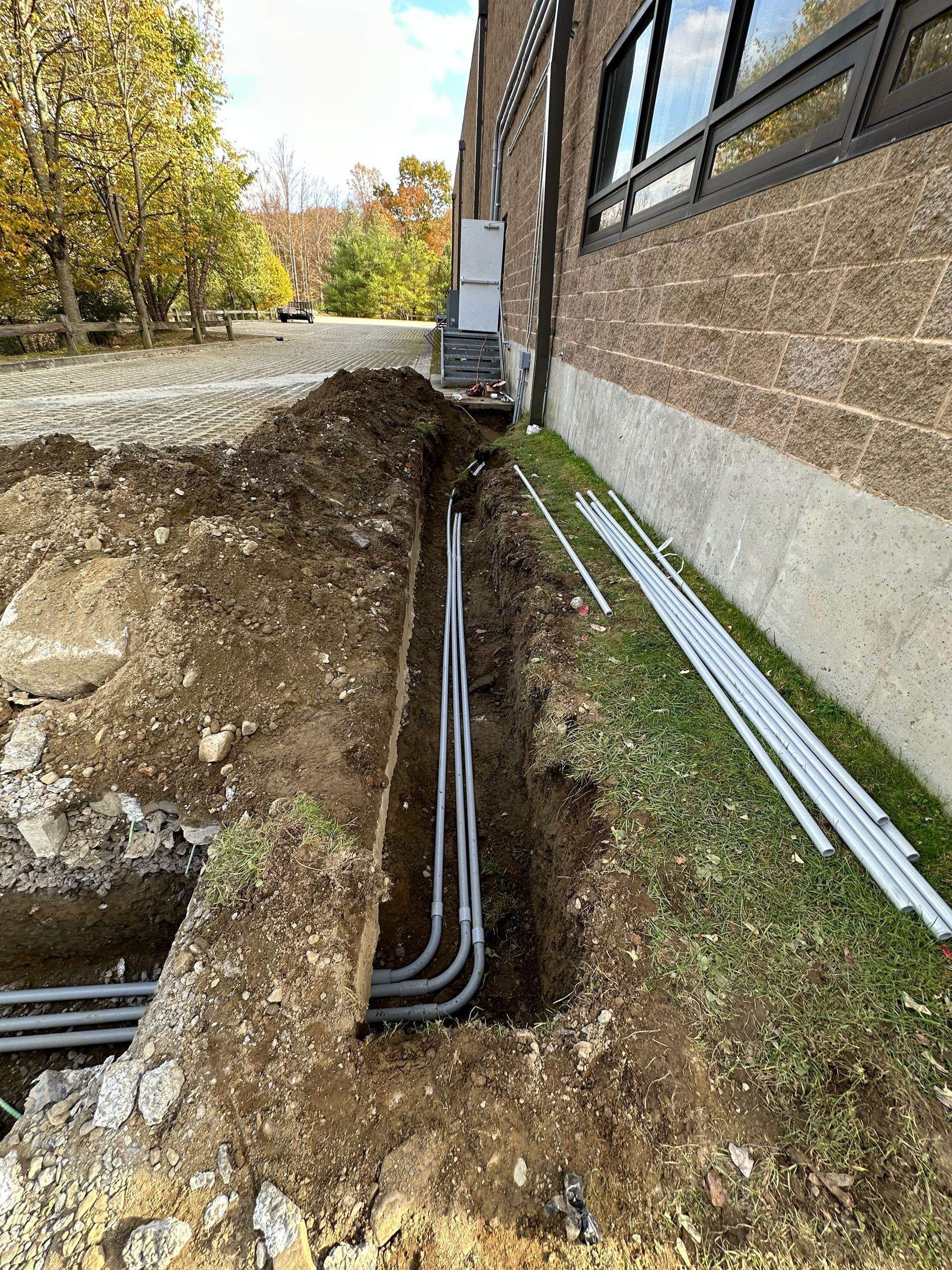 A trench alongside a brick building foundation containing several grey PVC electrical conduits, with pipes lying on grass.