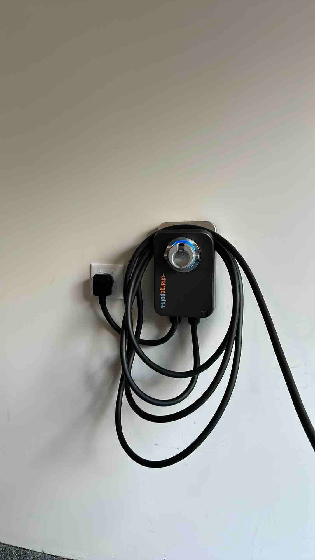 A wall-mounted black electric vehicle charger with a coiled cable plugged into a standard wall outlet.