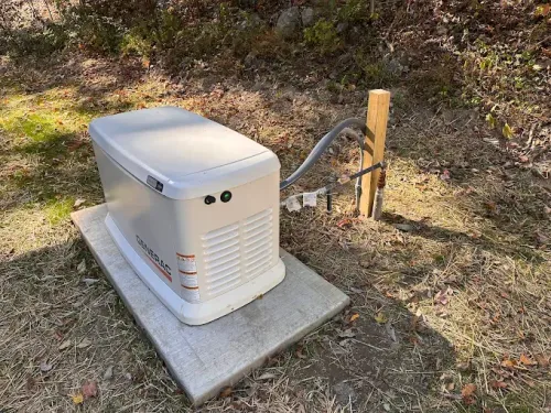 A white Generac standby generator sits on a concrete pad outdoors near a wooden post with utility conduits.