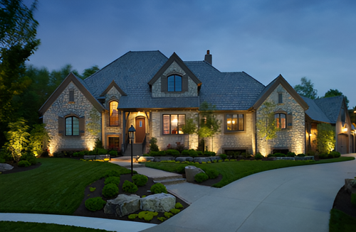 A stone luxury home at dusk with illuminated windows and landscape lighting, surrounded by a lush lawn and a driveway.