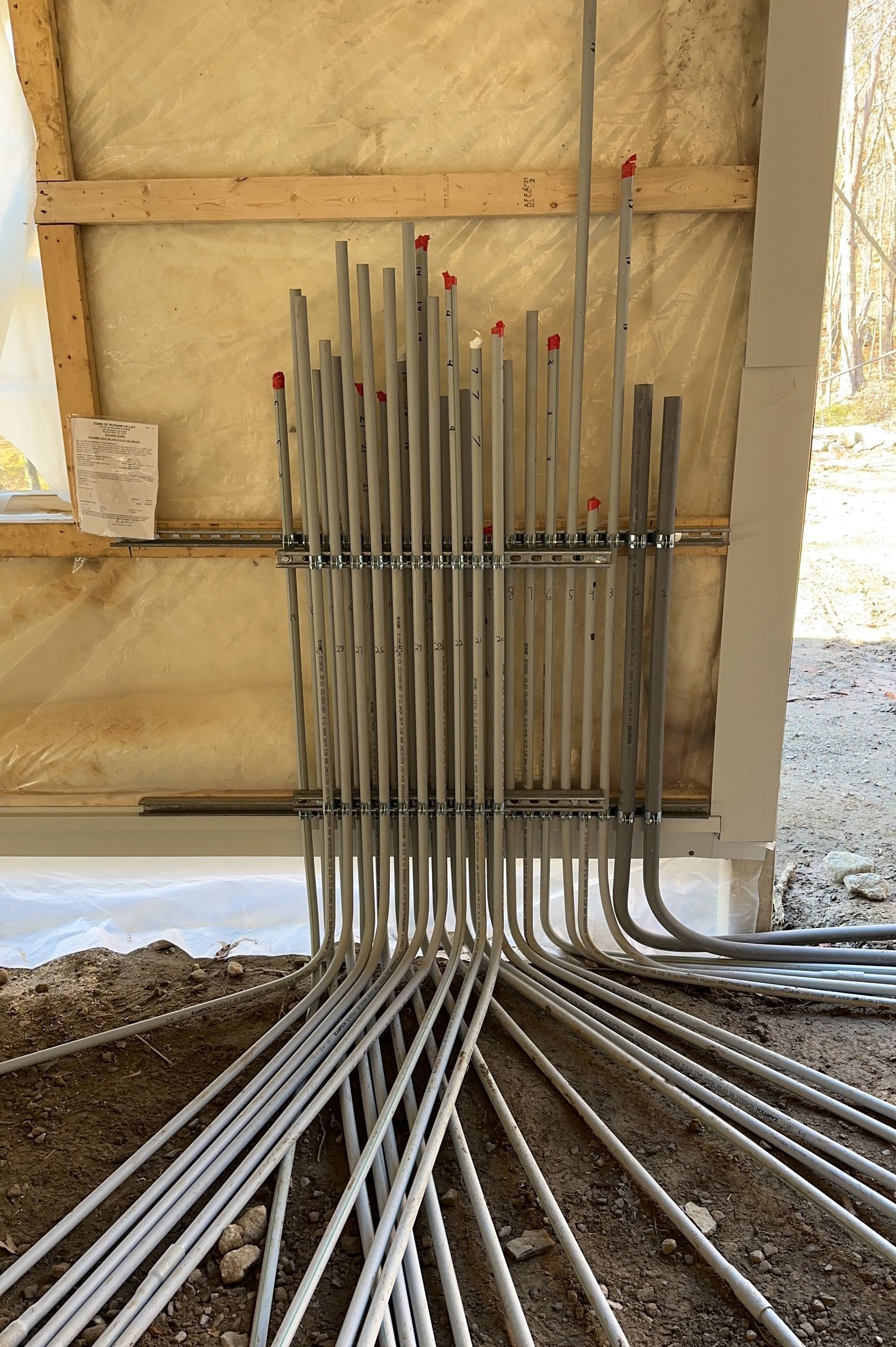 A collection of vertical grey electrical conduits mounted in a row on a wooden construction wall above a dirt ground.