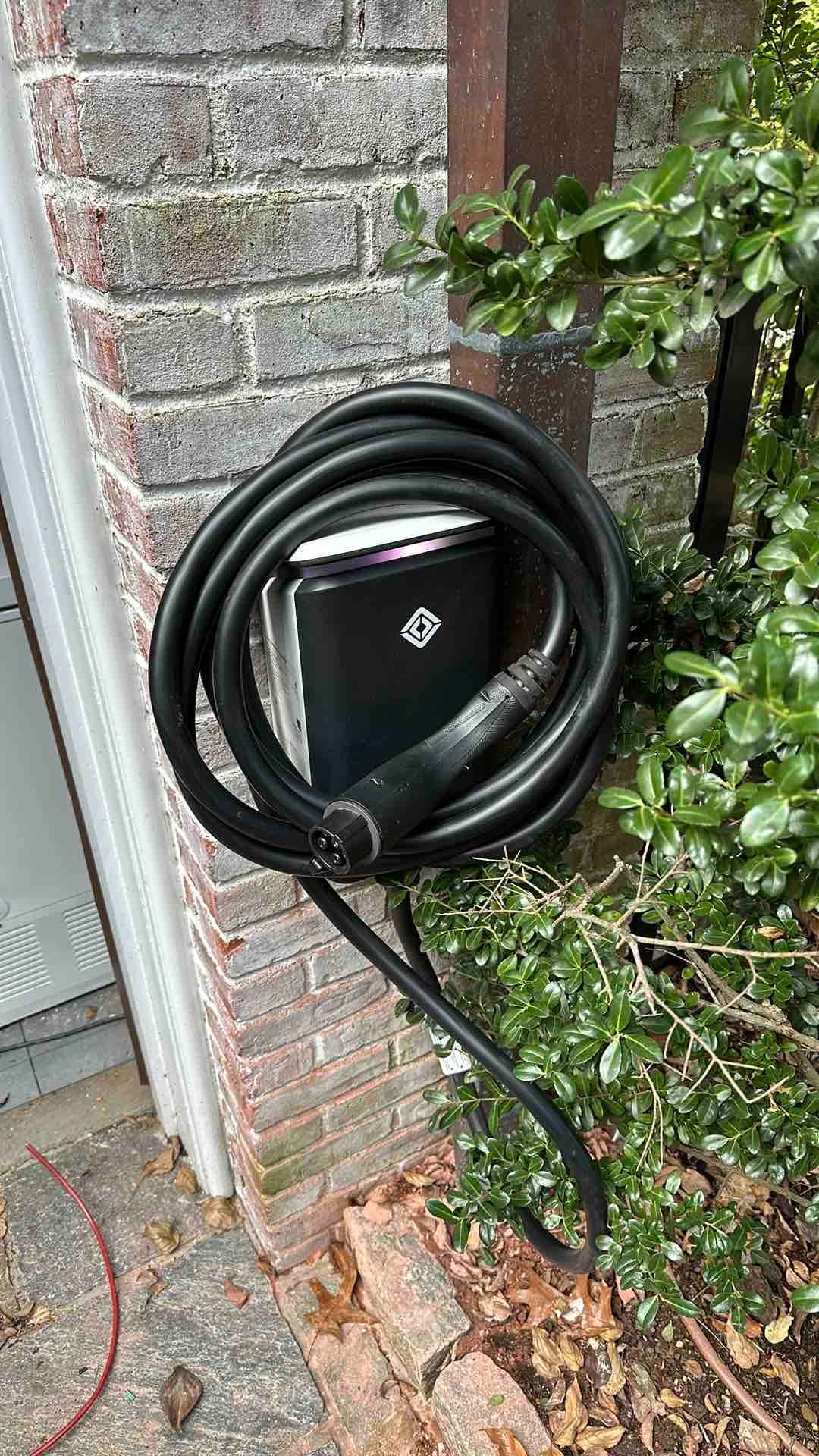 An electric vehicle charging station mounted on a brick wall outdoors, with a black cable coiled around it.
