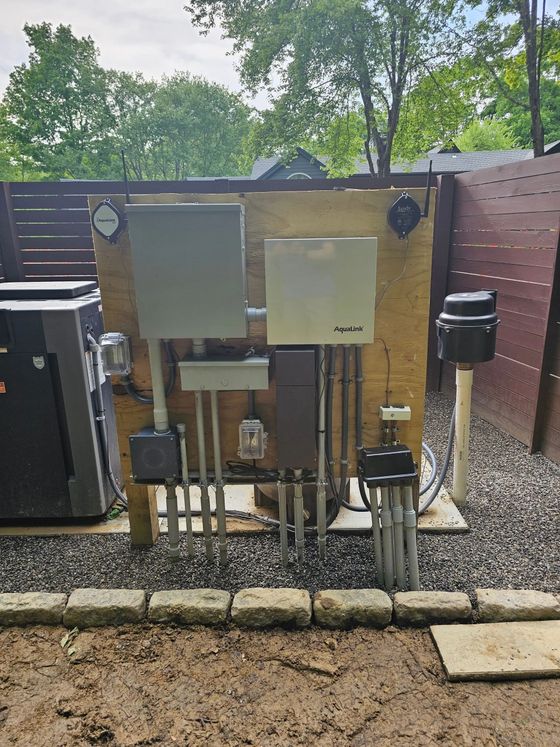 Electrical equipment, including a gray transfer switch and panel, mounted on a plywood board outdoors with conduit.