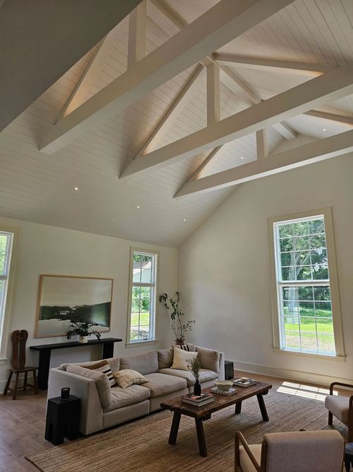 A bright living room with white vaulted ceilings, exposed wooden beams, a gray sectional sofa, and tall windows.