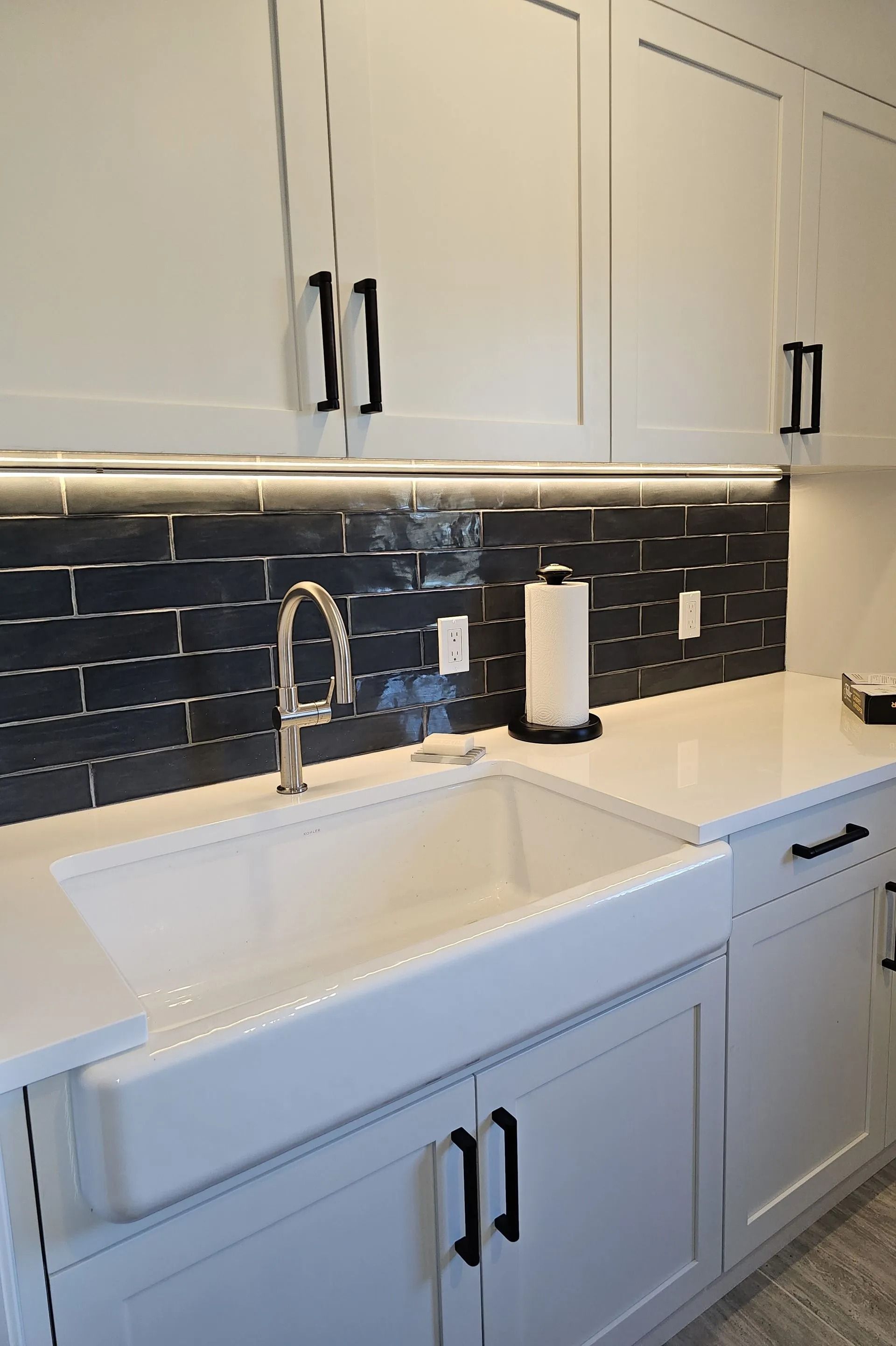 White farmhouse sink and cabinets with black hardware and a dark subway tile backsplash in a modern kitchen.