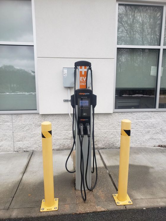 An electric vehicle charging station stands between two yellow bollards against a wall with windows.