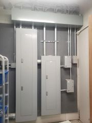 Two gray electrical panels mounted on a wall with metal conduits running upward into a horizontal wire trough.