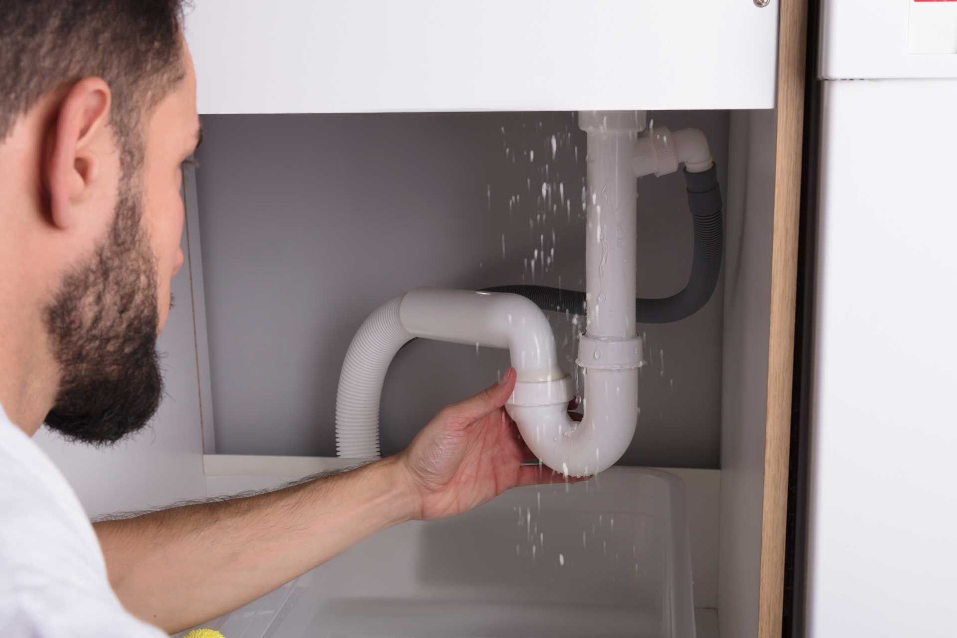 Man inspecting a leaking white sink drain pipe under a sink. Water is spraying. Man inspecting a leaking white sink drain pipe under a sink. Water is spraying.