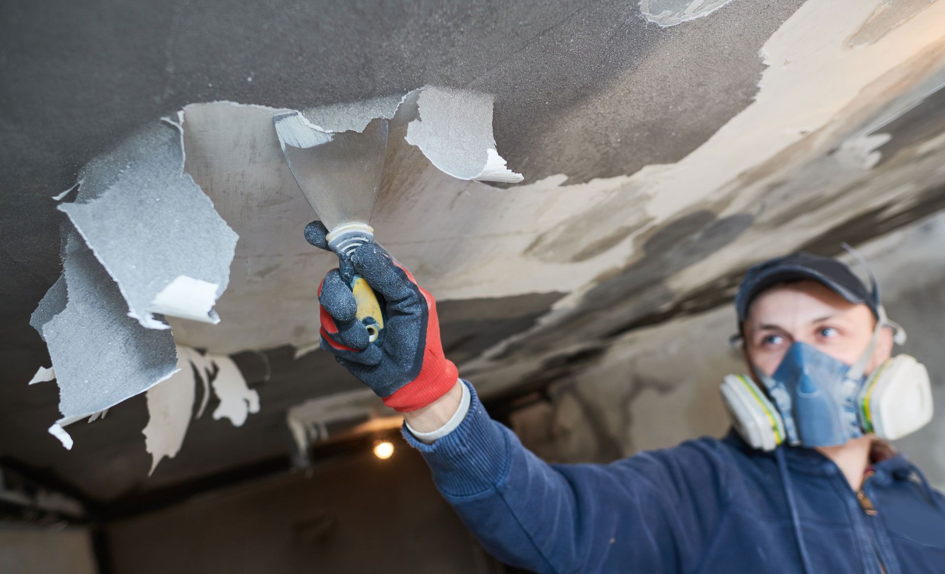 Person in mask removes peeling ceiling material with scraper. Interior, poor lighting. Person in mask removes peeling ceiling material with scraper. Interior, poor lighting.