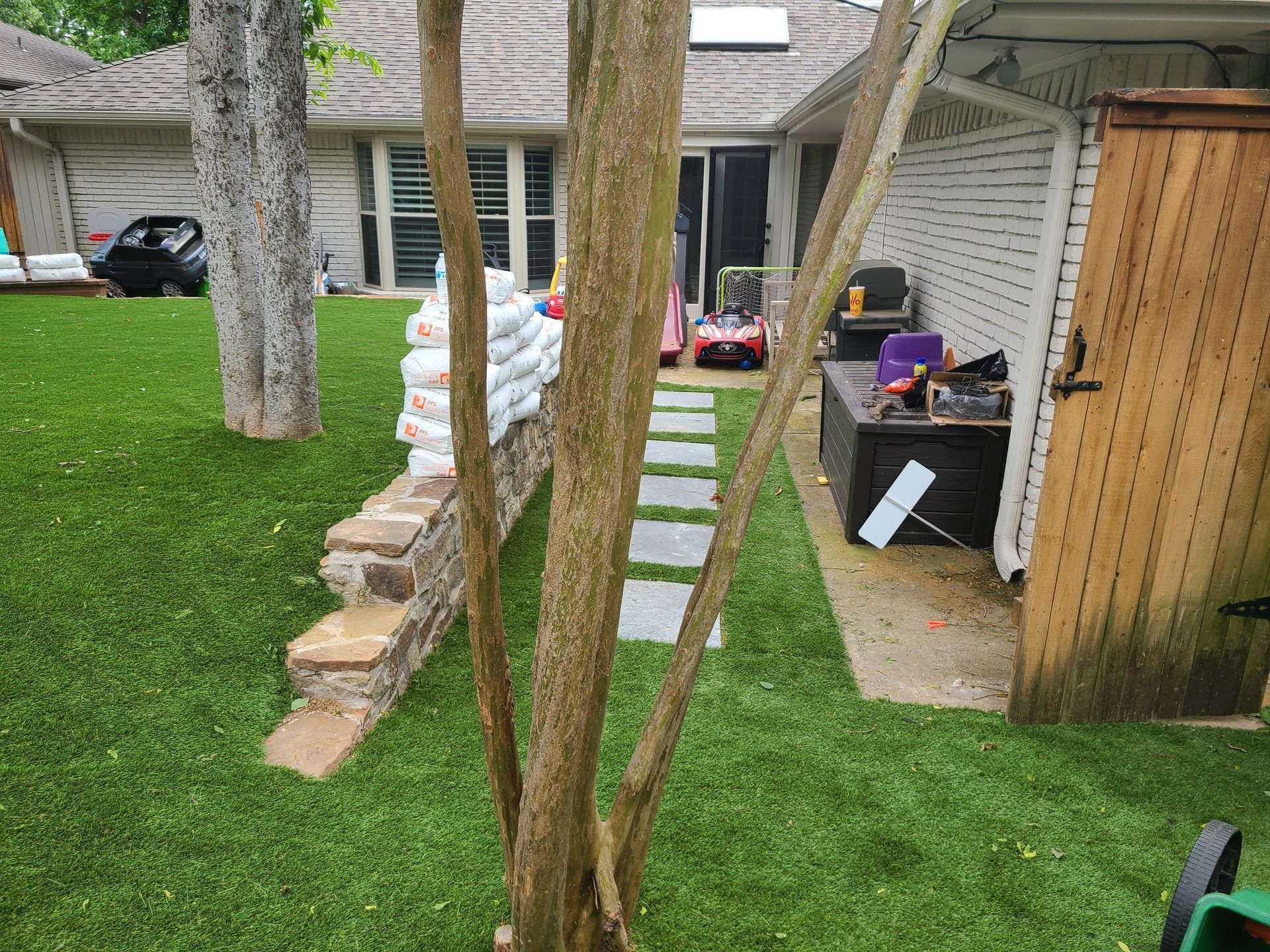 Green yard with a stone path, retaining wall, tree, and shed in the background.