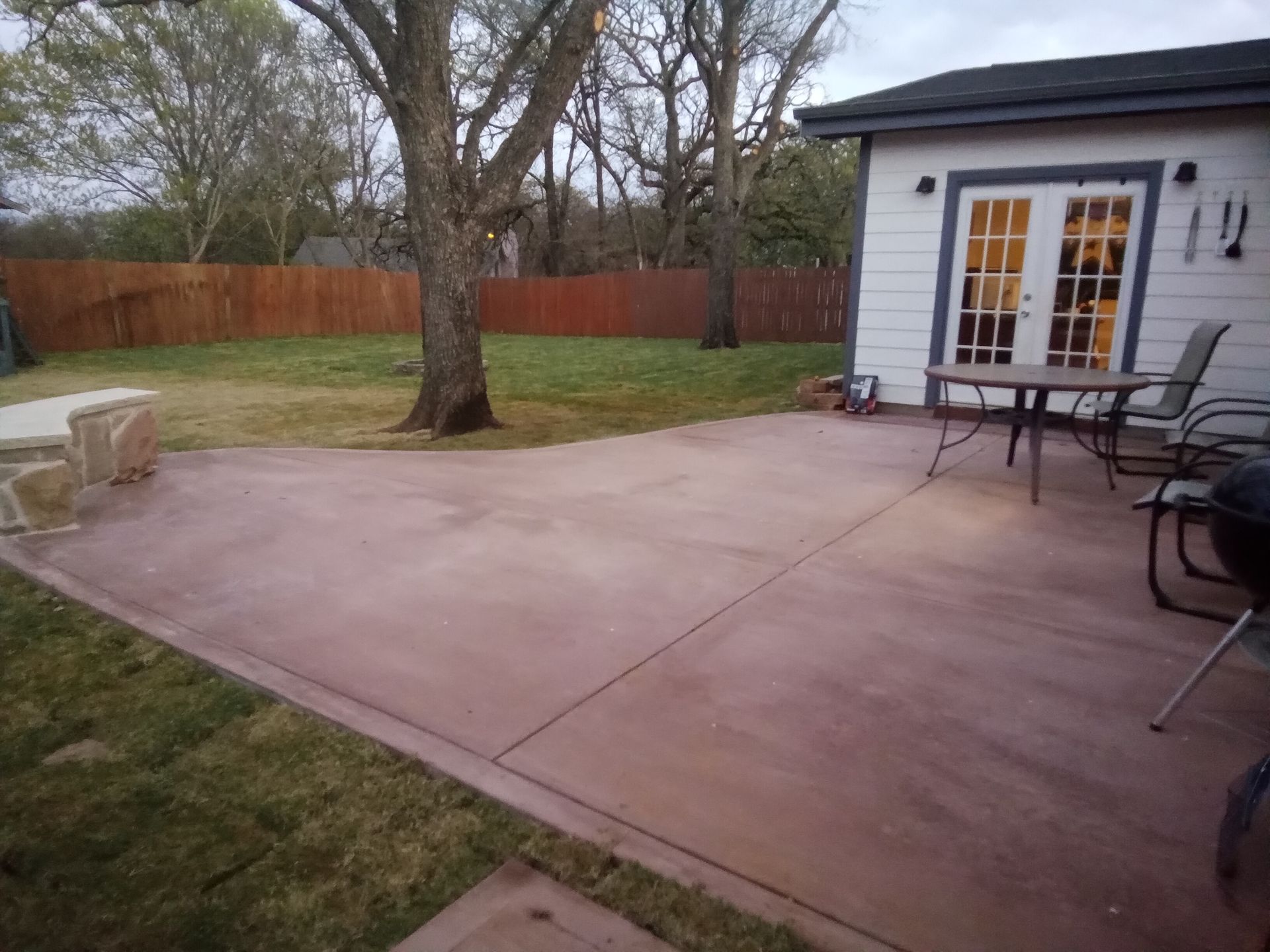 Backyard with concrete patio, table, chairs, and grill. Brown wooden fence in background, trees, and grass.