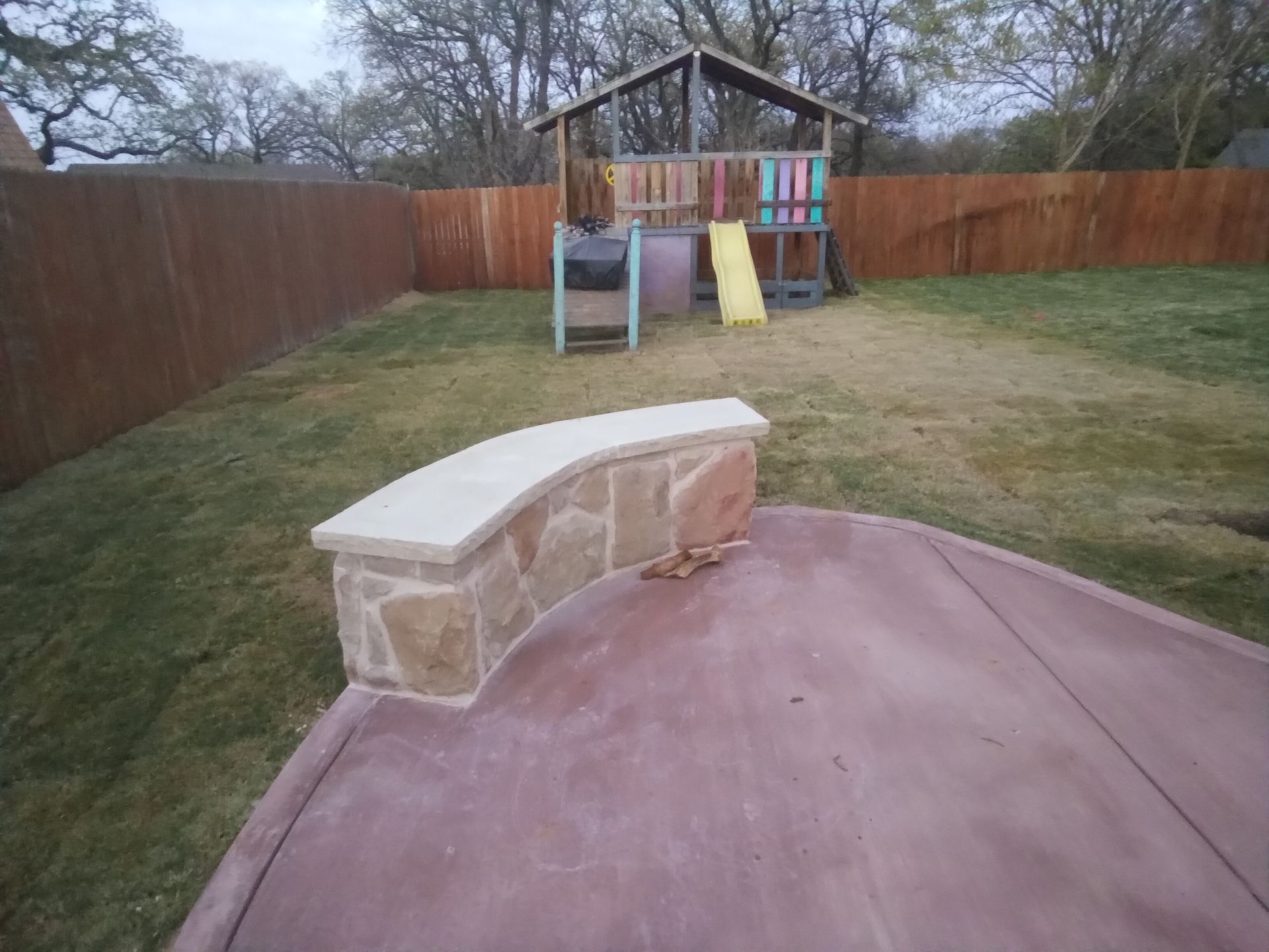 Backyard with stone wall, patio, playhouse, and brown fence.