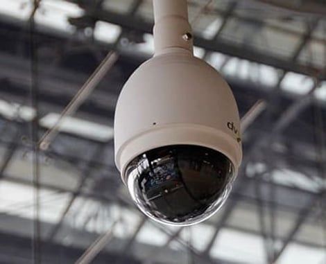 A white dome security camera hangs from a ceiling structure, set against a blurred background of architectural beams.