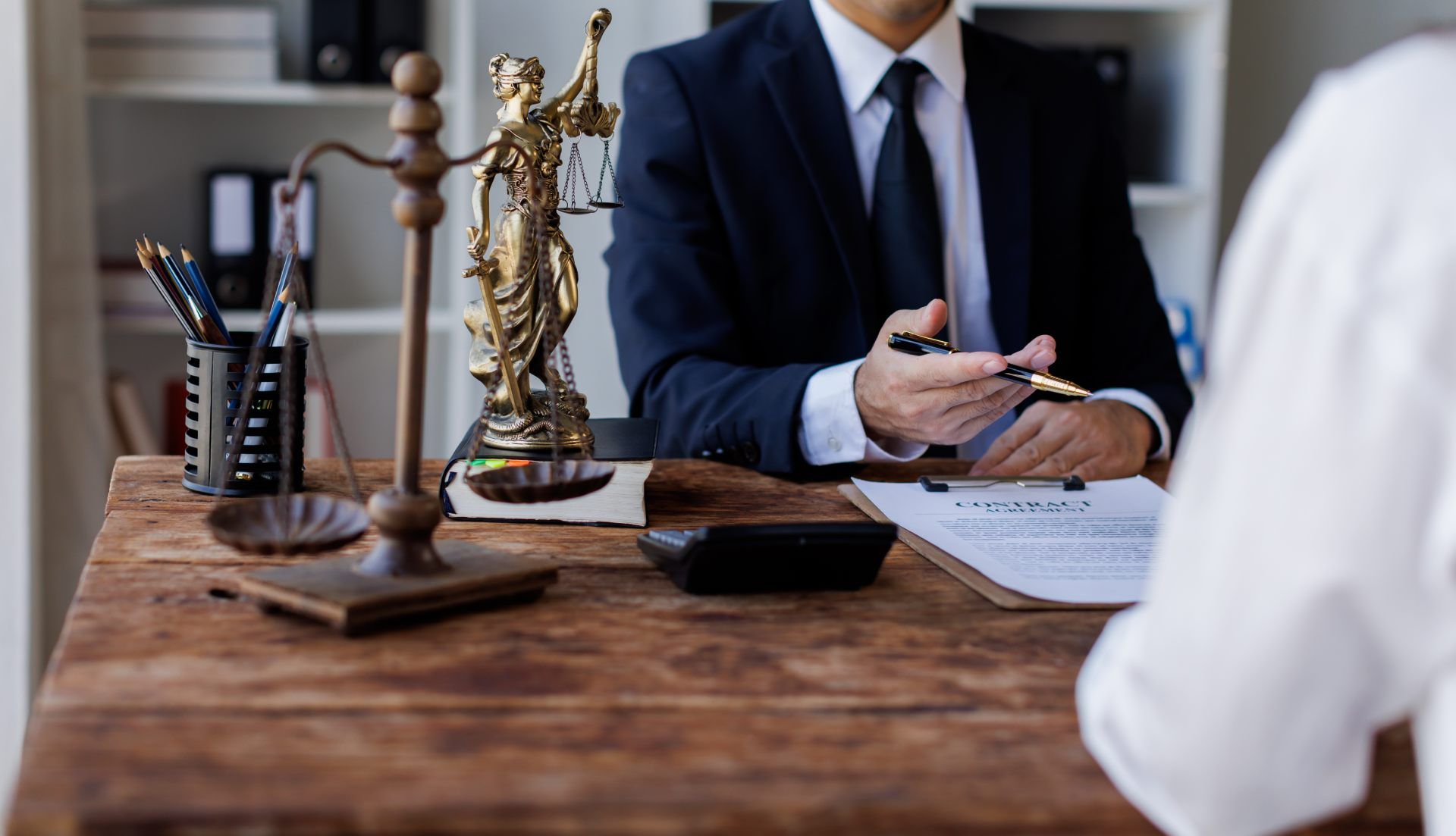 A professional in a suit gestures while discussing documents at a wooden desk with a Scales of Justice statue.
