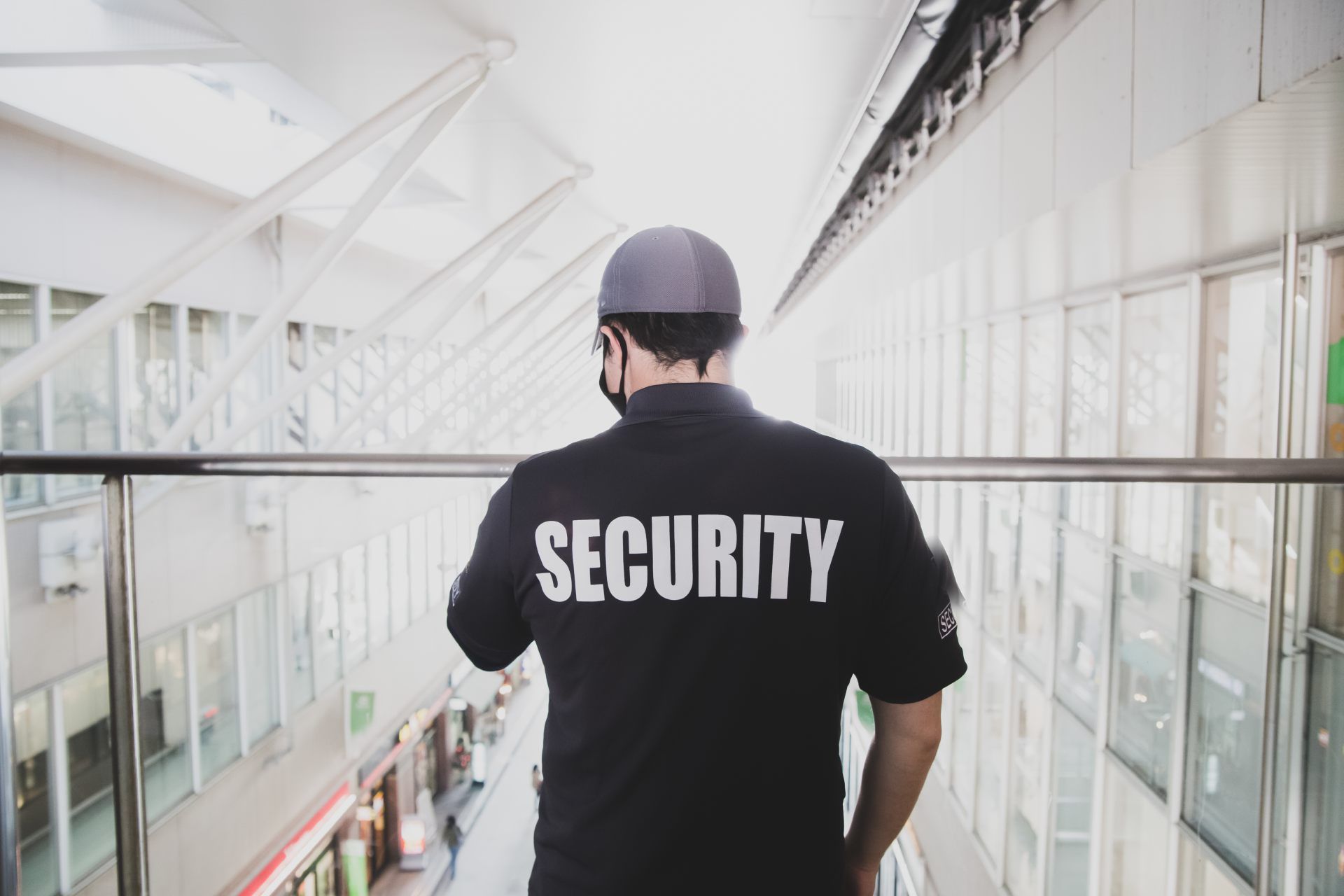 A security guard in a black uniform stands on a balcony overlooking a modern, brightly lit commercial corridor.