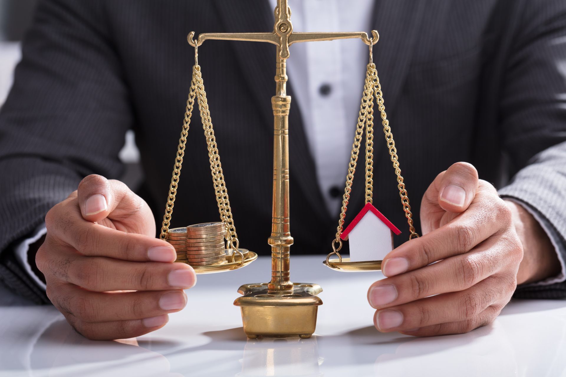 A person in a suit balances a stack of coins and a small model house on a brass scale.