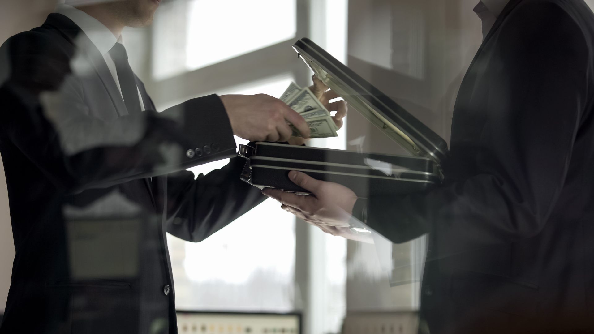 Two people in suits exchanging a stack of paper currency from an open briefcase.