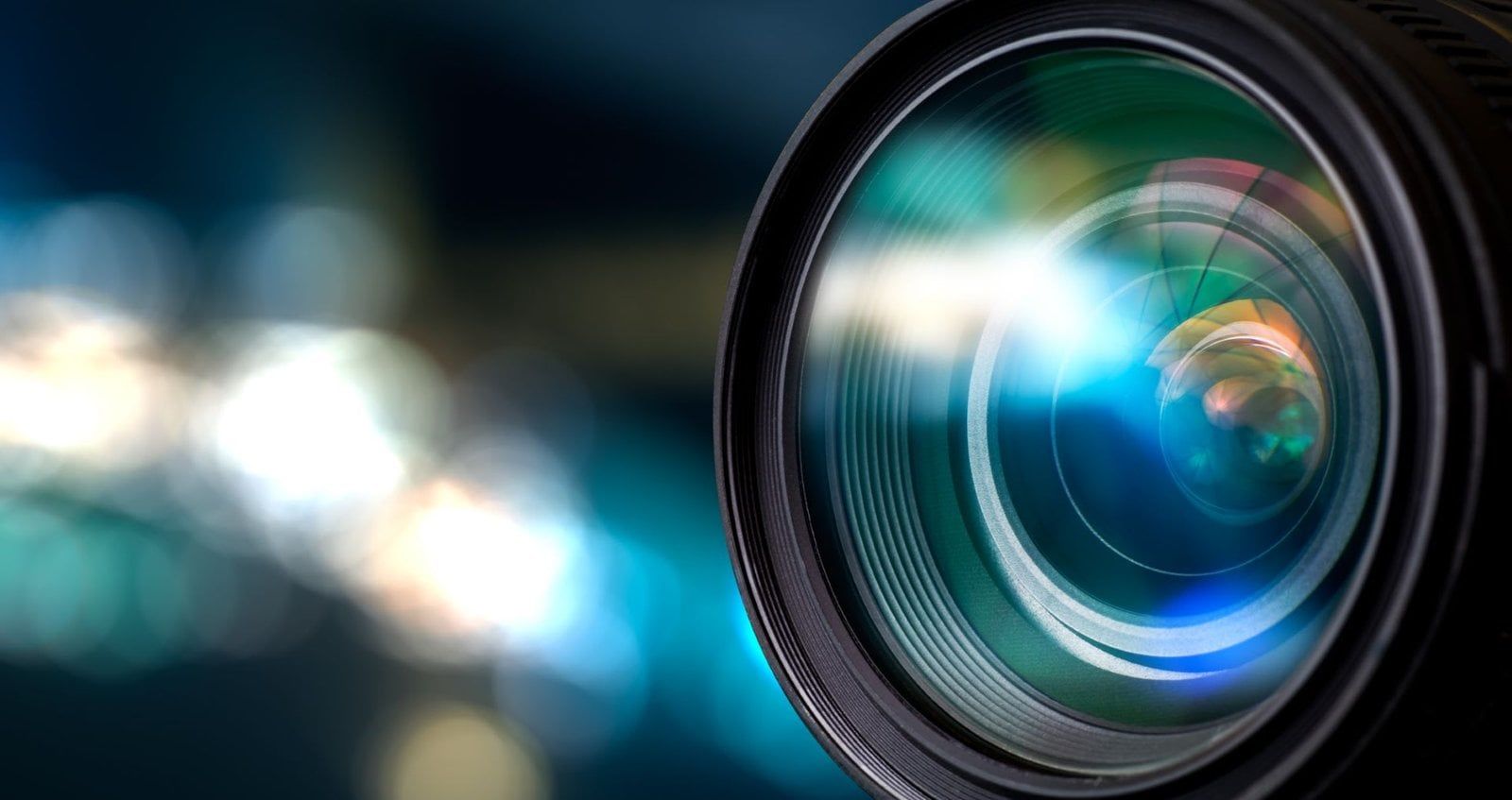 A close-up view of a camera lens with a blue light reflection, set against a blurred, dark background of bokeh lights.